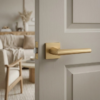 Minimalist square internal door handles in matt gold, showcasing the Jasmina lever on a square rosette against a pale grey internal door for a warm, contemporary finish.