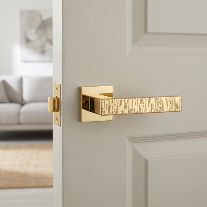 Amarylia - Polished Gold - White Door Amarylia on Square Rosette in polished gold, showcasing modern designer door handles with an intricate textured lever on a white interior door.